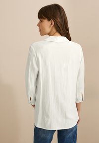 Camisa de tela arrugada blanca con cuello y mangas tres cuartos, que presenta una textura sutil y aberturas en los extremos de las mangas.