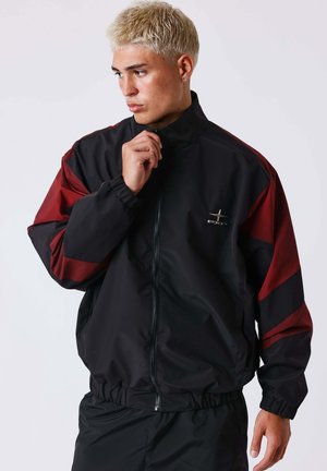 Giovane con capelli biondi corti che indossa una giacca con zip nera e rosso scuro, che tocca il colletto e guarda verso sinistra.