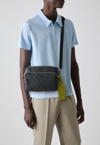 Bolso crossbody negro texturizado con cremallera plateada, detalle del logotipo y etiqueta brillante verde desmontable. Lleva puesto sobre una camiseta polo azul claro.