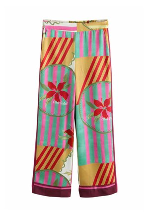 Person trägt bunte Hose mit pinken Blumen, grünen und gelben Streifen, steht auf Steinpflaster neben einem Metallgeländer mit Ozean im Hintergrund.