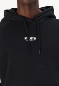 Čierna mikina s kapucňou a šnúrkami, s bielym logom "HOLLISTER" a textom "Originálne dizajny". Mäkkučká, štruktúrovaná tkanina.