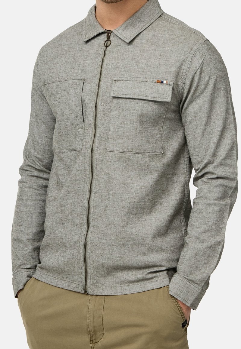 Homme portant une veste zippée gris clair avec deux poches poitrine, dont une avec rabat et petite étiquette colorée, associée à un pantalon beige.