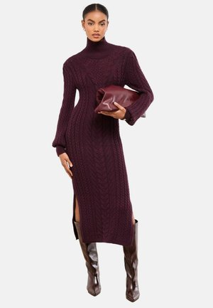 Stickad poloklänning i djupt vinrött med flätade mönster, sidosprund och ribbade muddar. Modellen håller en vinröd clutch.