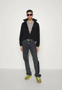 Jaunas vyras, dėvintis juodą striukę, pilką marškinėlį, tamsias džinsus, neoninės geltonos sportbačius, akinius nuo saulės ir karolius, stovi prieš vienodą foną.