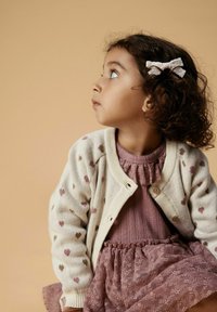 Jasno beżowy kardigan w serca, nałożony na różową sukienkę z prążkowanego materiału z falbaną przy dekolcie, w zestawie z akcesorium do włosów w kształcie kokardy.