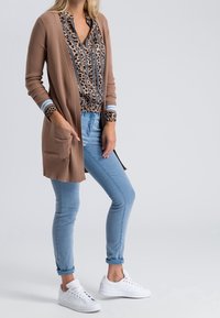 Bruine geribde cardigan met lange mouwen en zakken, gedragen over een blouse met luipaardprint en blauwe strepen. Lichtblauwe jeans en witte sneakers.