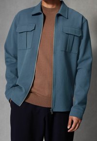 Lichtblauwe jas met ritssluiting en twee borstzakken over een bruine trui. De jas heeft een gestructureerde kraag en is gemaakt van een gestructureerde stof.