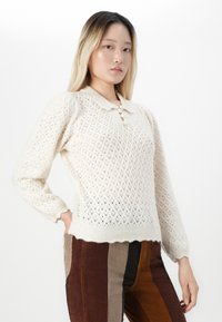 Junge Frau in einem cremefarbenen Strickpullover mit Kragen und mehrfarbigen senkrecht gestreiften Cordhosen, die vor einem weißen Hintergrund steht.