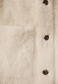 Manteau en fausse fourrure blanche avec une surface douce et texturée. Présente des boutons marron et un design épuré.