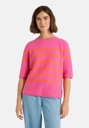 Frau mit schulterlangem dunklem Haar trägt ein pinkes Strickoberteil mit orangen horizontalen Streifen und hellblaue Jeans vor weißem Hintergrund.