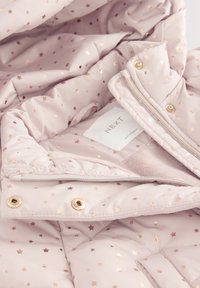 Intérieur de veste matelassée rose avec imprimé étoiles en or rose. Présente une texture lisse, une fermeture éclair et des accessoires de fixation en or. Étiquette d'entretien visible.