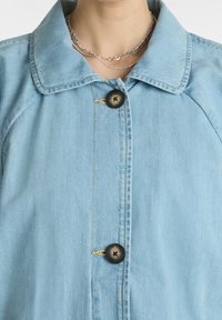 Vaaleansininen farkkutakki, jossa on klassinen kaulus, kaksi nappia ja hienovarainen saumadetalji etulistan kohdalla. Sileä rakenne.