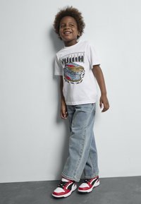 T-shirt blanc à manches courtes avec un imprimé graphique, associé à un jean ample bleu clair et des baskets rouges, noires et blanches.