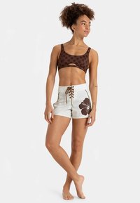 Barna virágmintás kivágott részlettel ellátott rövid felső, fehér rövidnadrággal, amely nagy barna hibiszkusz mintát és fűzős elülsőt visel.