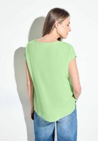 Top verde a maniche corte con scollo tondo e orlo curvo. Il tessuto appare leggero e morbido, abbinato a jeans blu.