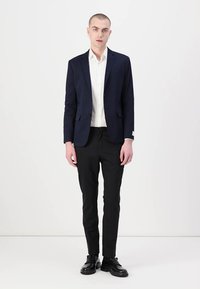 Marineblazer met notch-revers, wit overhemd en zwarte pantalon. Model draagt zwarte schoenen en staat tegen een effen witte achtergrond.