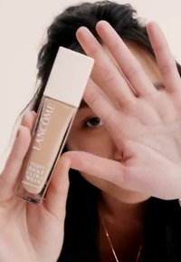 Bouteille de fond de teint Lancôme en verre transparent, liquide beige et capuchon blanc. Une main partiellement obstrue le visage, montrant une texture lisse et une teinte neutre.