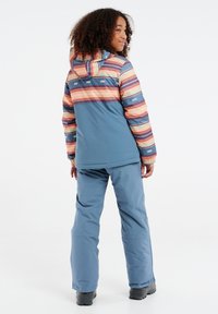 Blauwe skijas met een meerkleurig gestreept patroon, verstelbare capuchon, elastische mouwen. Gecombineerd met bijpassende blauwe skibroek en zwarte laarzen.