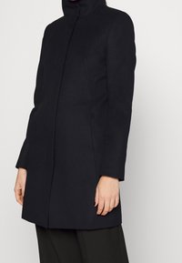 Nainen, jolla on yllään istuva musta takki, jossa on korkea kaulus, pitkät hihat ja piilotettu etulukko, yhdistettynä mustiin housuihin.