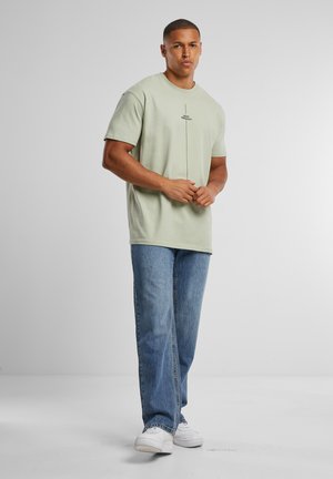 Homme debout, mains jointes, portant un t-shirt vert clair avec un petit texte, un jean bleu et des baskets blanches sur un fond uni.