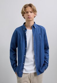 Joven con cabello rubio lleva una camisa de mezclilla azul desabotonada sobre una camiseta blanca y pantalones beiges, de pie contra un fondo claro y uniforme.