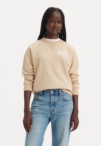 Sudadera beige de Levi's con un logo blanco, cuello redondo y puños de canalé, combinada con unos vaqueros de cintura alta en azul claro.