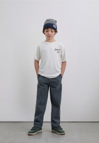 Bílé tričko s krátkým rukávem s tištěným logem, šedé manšestrové kalhoty a šedo-modrá čepice. Obuv má tmavě zelené a hnědé akcenty.