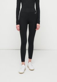 Oseba v črnih visokih pajkicah in belih supergah stoji pred enobarvno belo ozadje.