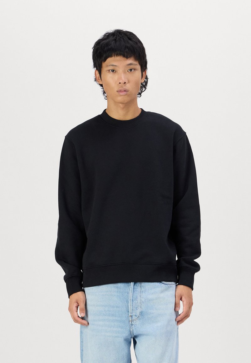 Black sweatshirt with ribbed neckline and cuffs, featuring a relaxed fit. Worn with light blue jeans, displayed against a plain background.