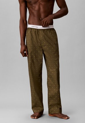 Homme torse nu portant un pantalon de pyjama Calvin Klein vert olive avec un petit motif de logo jaune, debout pieds nus sur un fond gris uni.