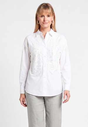 Femme aux cheveux blonds raides portant des boucles d'oreilles créoles, une chemise blanche brodée à boutons et un pantalon gris clair, debout devant un fond blanc.