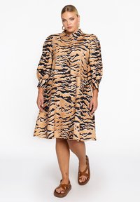 Tan-shirtjurk met zwart en bruin tijgerstrepen, ballonmouwen en een kraag, gecombineerd met bruine sandalen. Glad stofoppervlak.