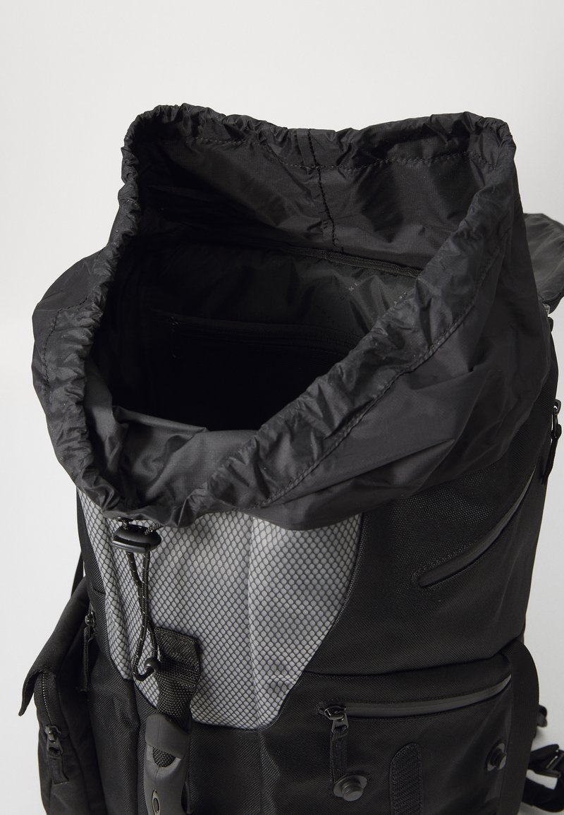 Sac à dos noir avec un haut resserré, doté d'un panneau gris texturé, de plusieurs poches et d'un tissu durable. À l'intérieur, un compartiment spacieux.