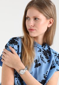 Silberne Uhr mit rundem Ziffernblatt und Metallband, am Handgelenk getragen. Die Person trägt ein blaues Hemd mit schwarzen Blumenmustern.