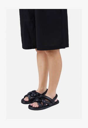 Zwarte gevormde sandalen met dikke gevlochten banden, platte zolen en open tenen, gemaakt van een soepel, flexibel materiaal. Gedragen met zwarte shorts.
