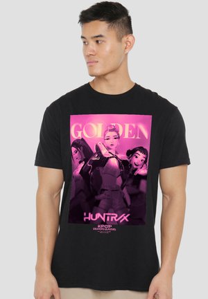 Man wearing black t-shirt featuring three animated female characters with "GOLDEN HUNTRX KPOP DEMON HUNTERS" text in pink and purple tones.