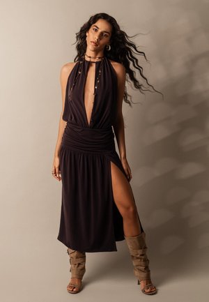 Femeie cu păr lung și ondulat, purtând o rochie fără mâneci, de culoare închisă, cu decolteu tip halter și despicătură frontală, precum și cizme maro, cu vârful deschis, stând în picioare pe un fundal neutru.