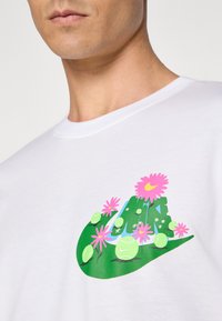 Camiseta blanca con un diseño gráfico verde de una colina con flores rosas y pelotas de tenis, mostrando colores vivos y formas juguetonas.