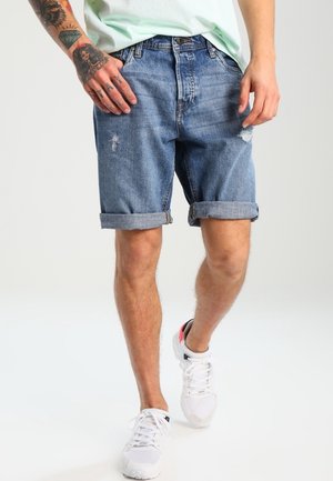 Personne portant un short en jean bleu retroussé avec de légères déchirures, une chemise claire, des baskets blanches et des tatouages visibles sur la main et le bras gauche.
