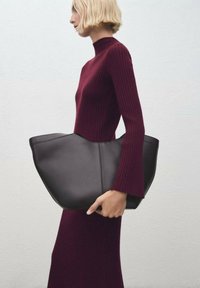 Borsa tote in pelle nera con una forma ampia e angolare e un design senza cuciture. Il modello indossa un vestito borgogna a coste, con maniche svasate.