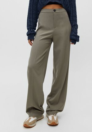 Person trägt lockere graue Hose, beige und weiße Sneaker und einen marineblauen, kabelgestrickten Pullover mit langen Ärmeln.