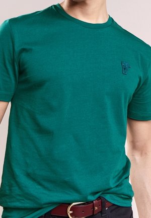 Homme portant un t-shirt vert sarcelle uni à manches courtes avec un petit logo brodé sur la poitrine et une ceinture marron avec une boucle dorée.
