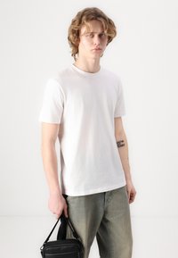 T-shirt bianca in cotone con scollatura rotonda, maniche corte e vestibilità comoda, abbinata a jeans verde chiaro e una piccola borsa nera.