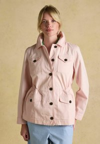 Lyshåret kvinde iført en lyserød knappet jakke med fire forlommer og blå-hvide stribede bukser mod beige baggrund.