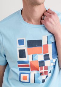 Light blue cotton t-shirt featuring a multicolored abstract geometric print in orange, navy, white, and blue with varying shapes and sizes.