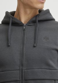 Siva zip-up dukserica od teksturirane tkanine, koja ima kapuljaču, vezice i mali logotip na prsima.