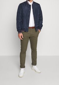 Uomo con giacca bomber blu navy, camicia bianca con bottoni, pantaloni verde oliva arrotolati, cintura marrone, calzini bianchi e sneakers bianche.