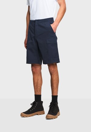 Mann trägt marineblaue Cargo-Shorts, schwarze High-Top-Sneaker mit beige Sohlen, schwarze Socken und ein weißes T-Shirt, steht mit einer Hand in der Tasche.