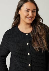 Zwarte gebreide cardigan met een gestructureerd ontwerp, ronde halslijn en gouden knopen langs de voorkant. Zachte, middelzware stof.