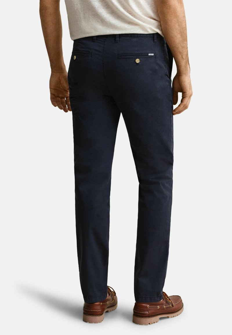 Hombre vestido con pantalones ajustados azul marino y zapatos de cuero marrón, posando contra un fondo claro y liso.